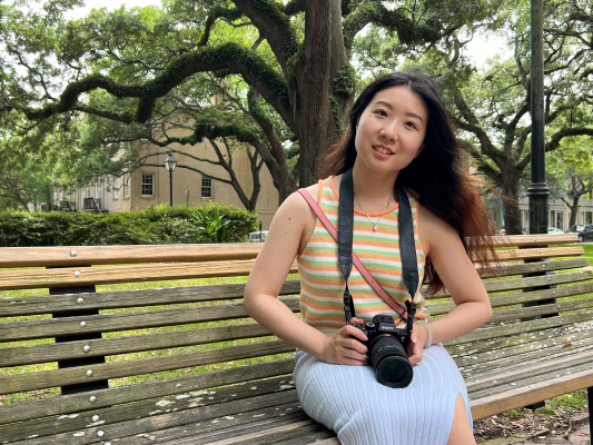 Kexin Song outdoors beneath a large tree, holding a camera
